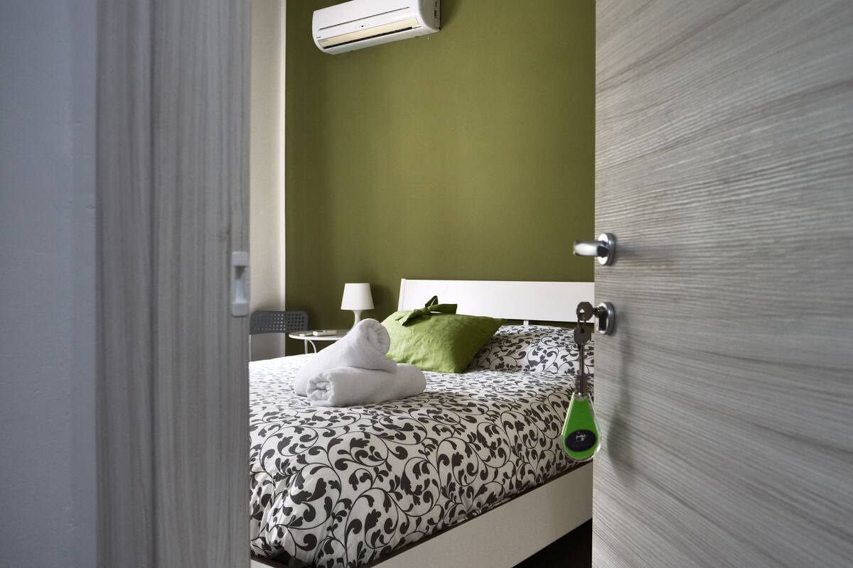 A bedroom with a bed, a green pillow, a white towel, a bedside table with a lamp, and a wall-mounted air conditioner.