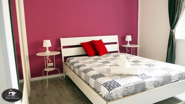 Una cama bien hecha con una colcha estampada, dos almohadas rojas y un cabecero blanco. Hay dos mesitas de noche con lámparas, y una cortina está parcialmente abierta.