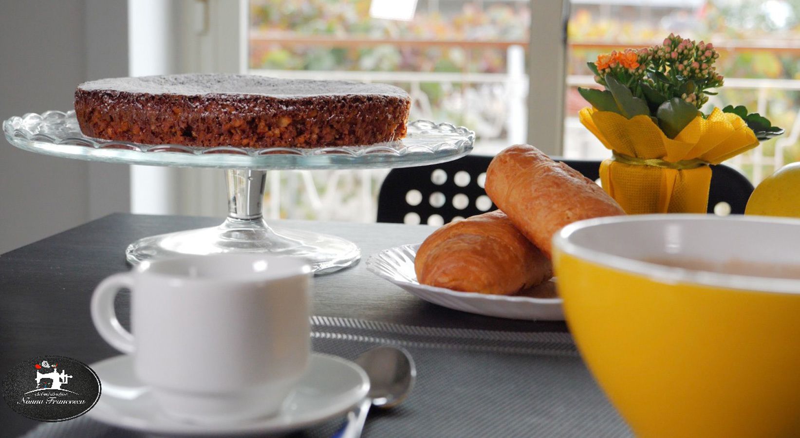 A breakfast setting with a cake on a glass stand, croissants on a plate, a yellow bowl, and a white cup on a table.