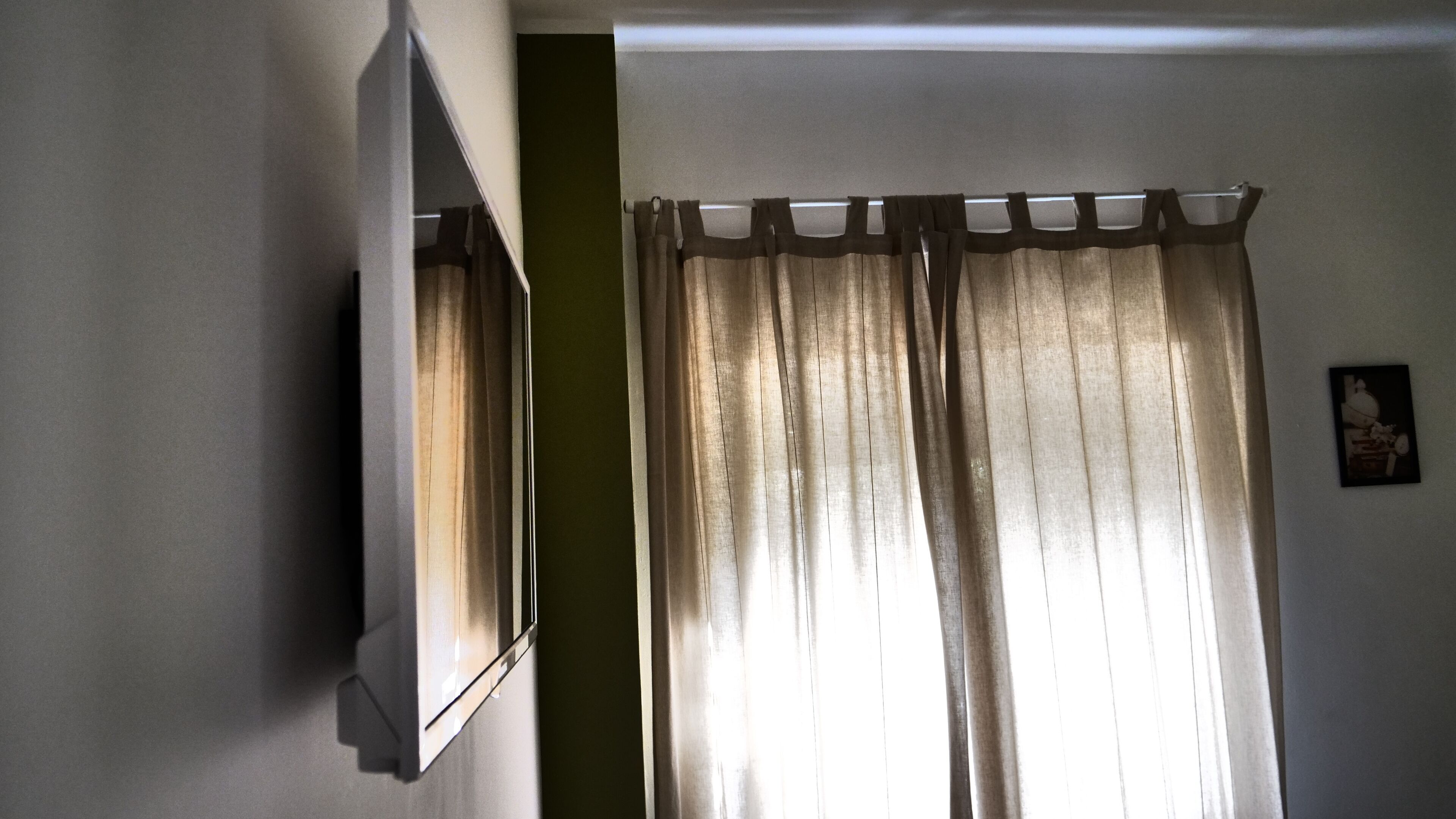 Une pièce dotée d’un téléviseur à écran plat fixé au mur, d’une fenêtre avec des rideaux beiges et d’un tableau encadré accroché au mur.