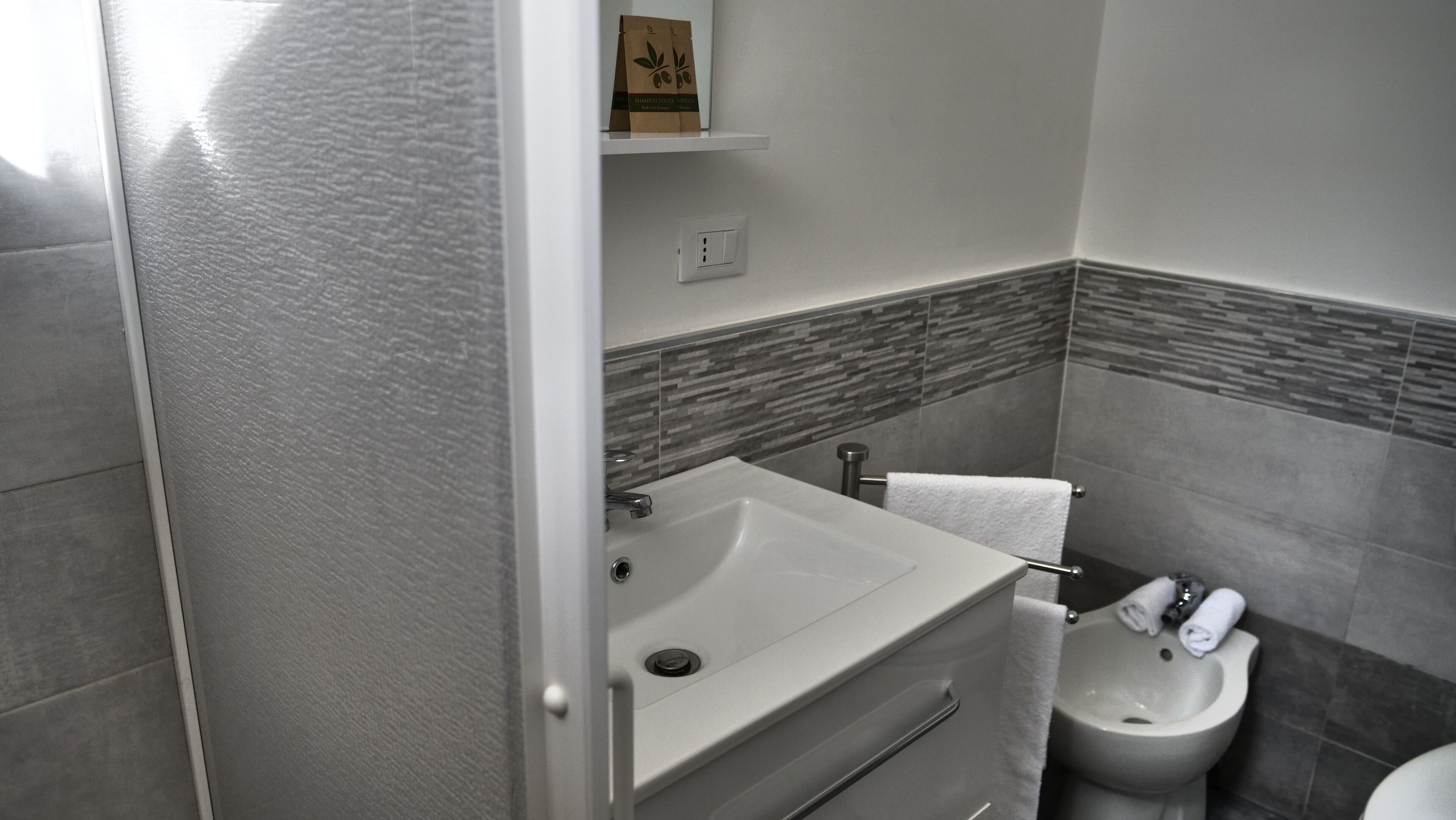 A bathroom with a white sink, a toilet, and a bidet. There is a towel rack above the sink and a small shelf with a bag of toiletries.