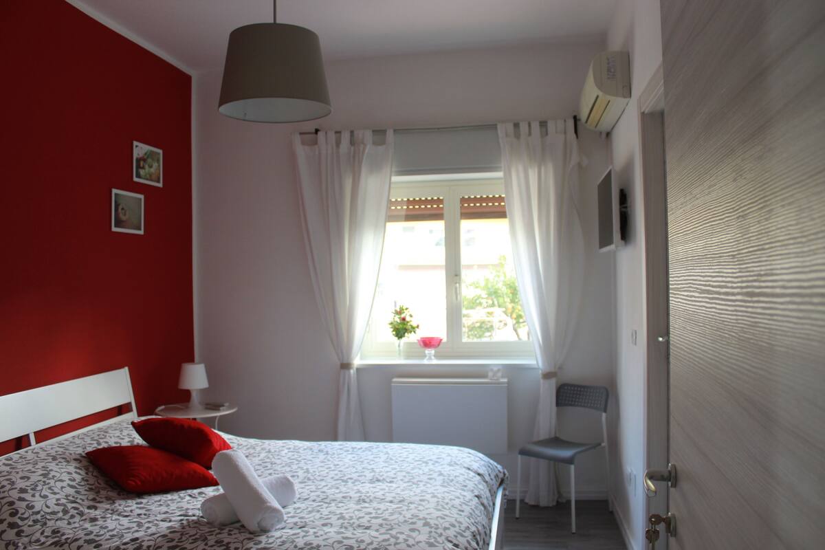 A bedroom with a bed, a window with curtains, a chair, and a radiator.