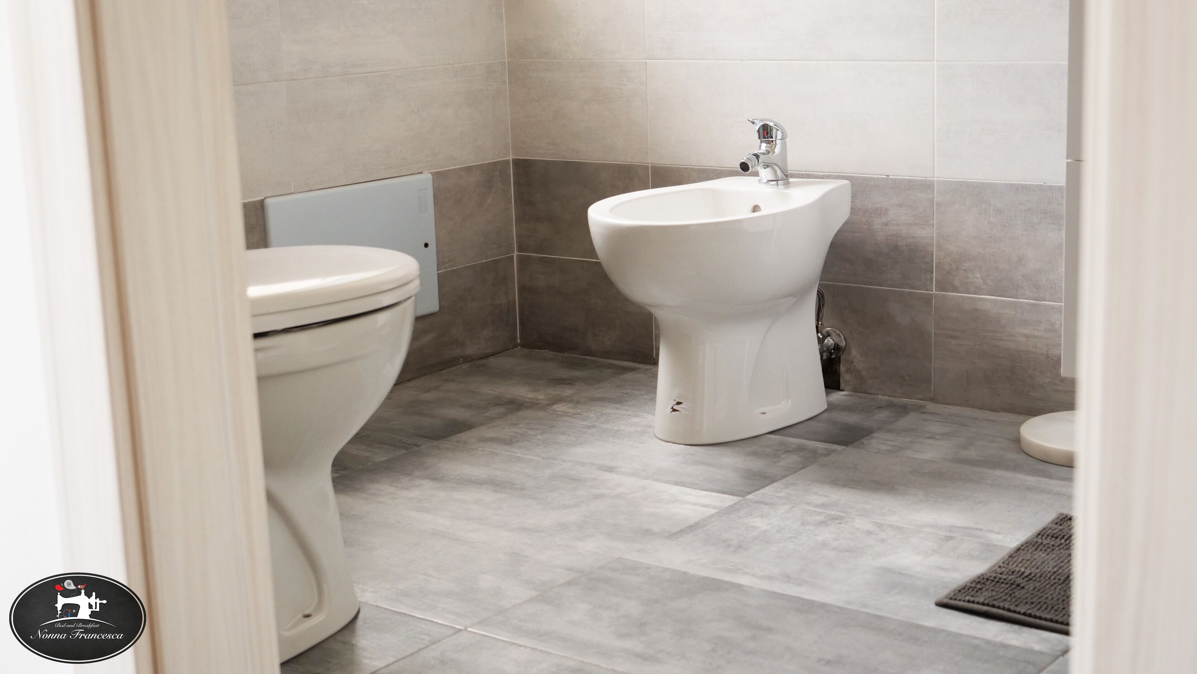 Ein Badezimmer mit Toilette und Bidet, gefliestem Boden und einem sichtbaren Logo an der Wand.