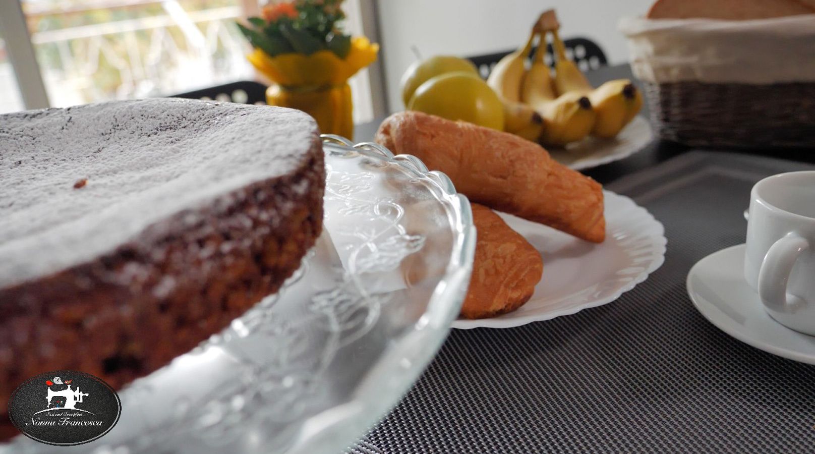 A cake on a glass stand, a plate with croissants, a cup, and a basket of bananas on a table.