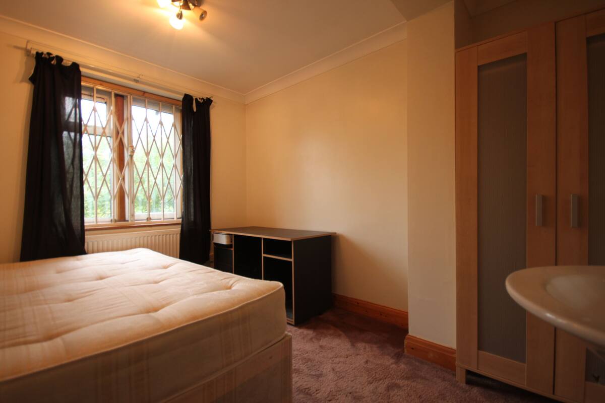 A bedroom with a bed, a window with curtains, a radiator, a wardrobe, and a sink.