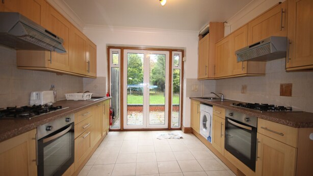 A kitchen with wooden cabinets, a gas stove, an oven, a washing machine, and a large sliding glass door leading to a garden. - Draycott Guest House (Harrow)
