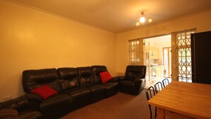 A living room with a black leather sofa, a wooden dining table, and a kitchen visible through an open door. - Draycott Guest House (Harrow)