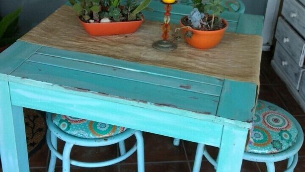 A turquoise-painted table with two chairs, each with a colorful cushion, and three potted plants on the table.