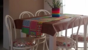 A dining area with a table covered by a colorful tablecloth, surrounded by white chairs with floral upholstery. A plant is placed on the table, and a window with curtains is visible in the background.