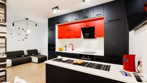 Une cuisine moderne avec des meubles de couleur noire et rouge, un îlot central doté d’une machine à café et un coin salon avec un canapé et des fauteuils.