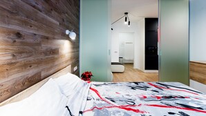 Une chambre moderne avec une tête de lit en bois, un lit recouvert d’une couvre-lit à motifs, une table de chevet avec une lampe, et une vue sur une pièce adjacente comprenant un canapé et un coin repas.