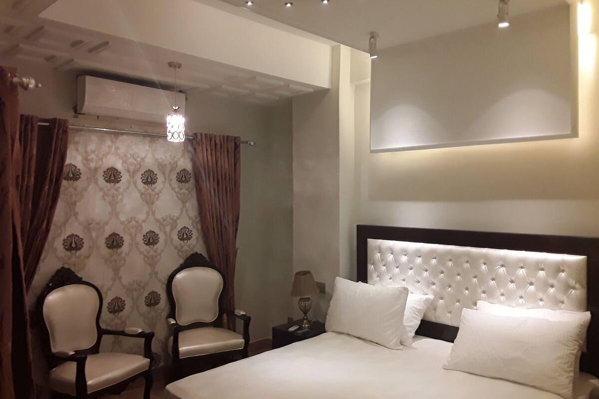 A hotel room with a large bed, two armchairs, a nightstand, and a wall-mounted air conditioning unit.