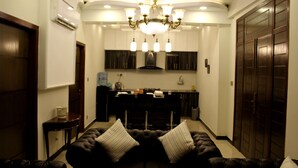 A living room with a dark brown sofa, two white striped pillows, a chandelier, and a kitchen in the background. - luxes Inn (Rawalpindi)