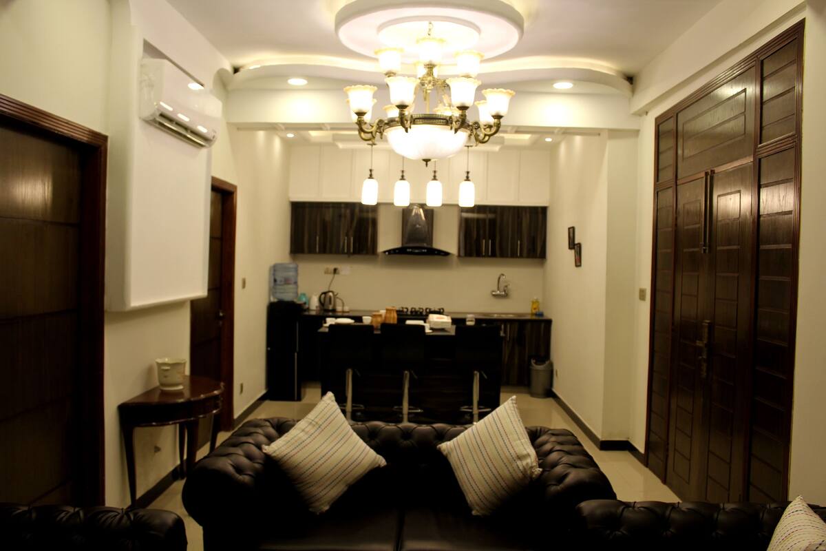A living room with a dark brown sofa, two white striped pillows, a chandelier, and a kitchen in the background.