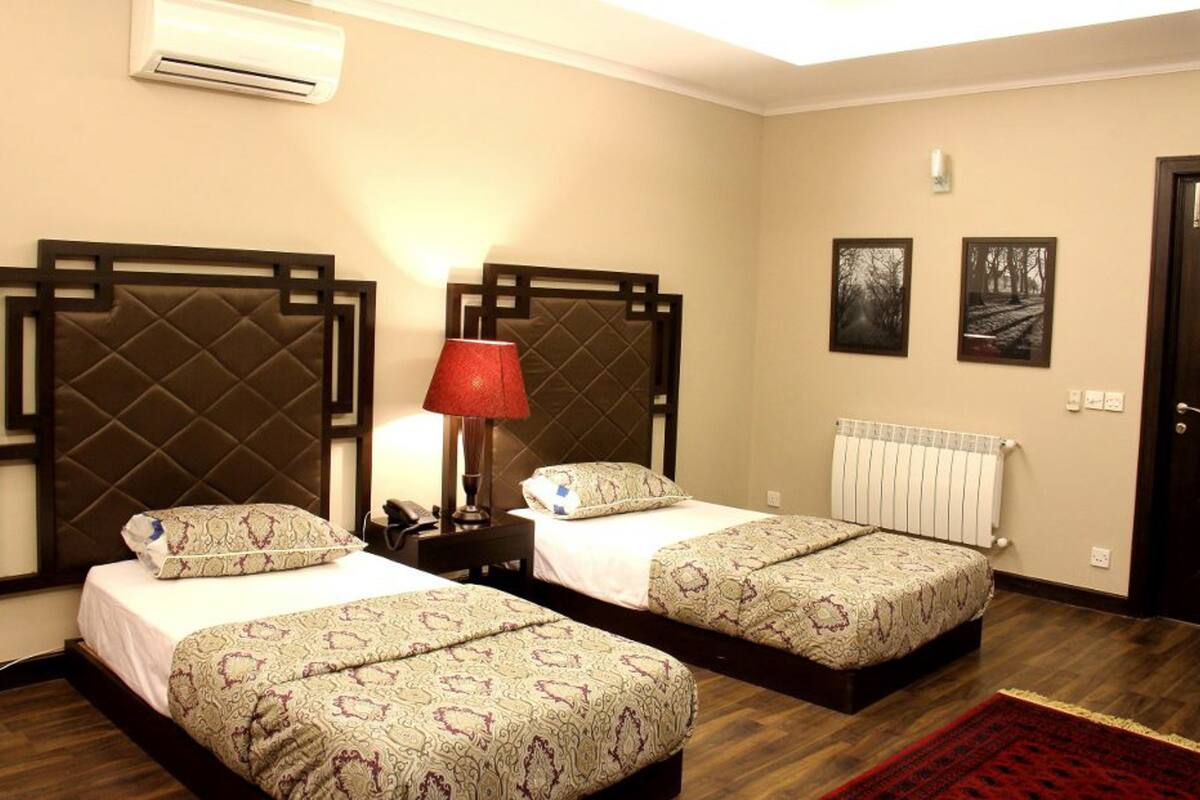 A hotel room with two beds, a red lamp, a radiator, and two framed pictures on the wall.
