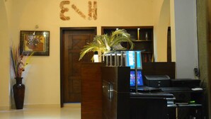 Un área de recepción con un escritorio de madera, computadora y una planta. Se ve una puerta al fondo y un cuadro en la pared.