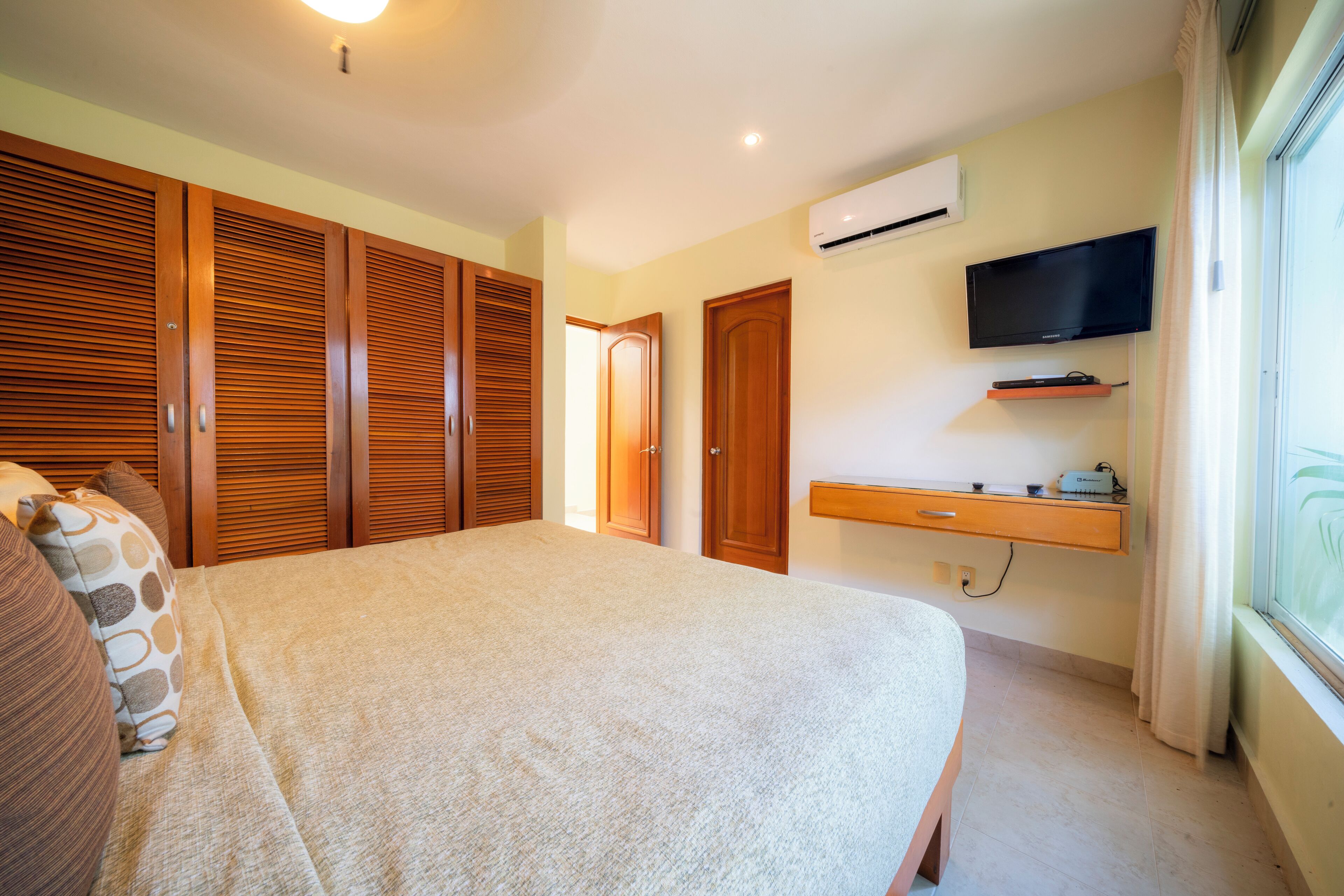 A bedroom with a bed, wooden wardrobe, a TV mounted on the wall, and an air conditioning unit.