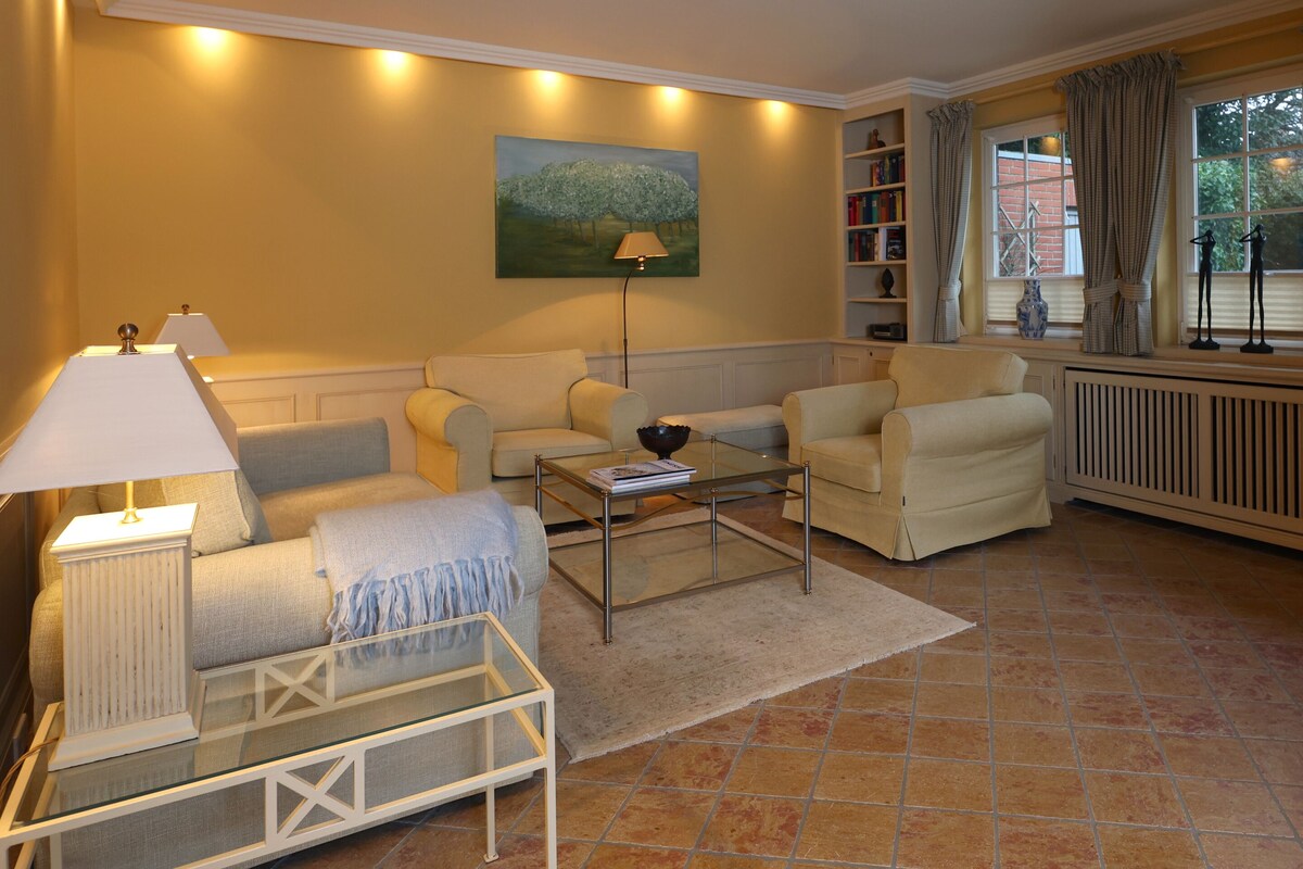 Una sala de estar con un sofá, sillones, una mesa de centro y una lámpara. Hay un cuadro en la pared y una estantería con libros.