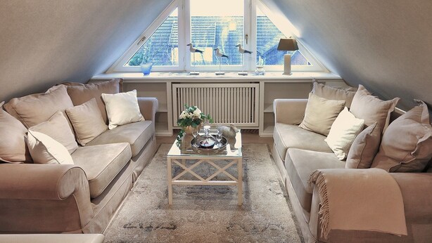 A cozy loft bedroom with a sloped ceiling, a large skylight, two beige sofas, a small coffee table, and a radiator. - Ferienwohnung Biberhüs OG (Sylt)