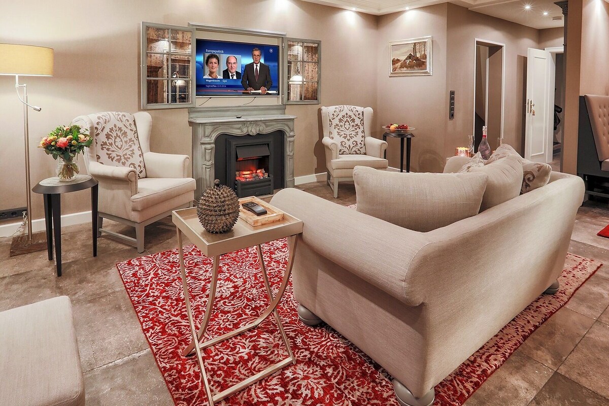 A living room with a fireplace, two armchairs, a sofa, a TV mounted above the fireplace, and a red patterned rug.