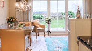 A dining area with a table set for two, a chandelier, and a view through large windows. - Ferienwohnung Sönshörn 8 Rechts (Sylt)