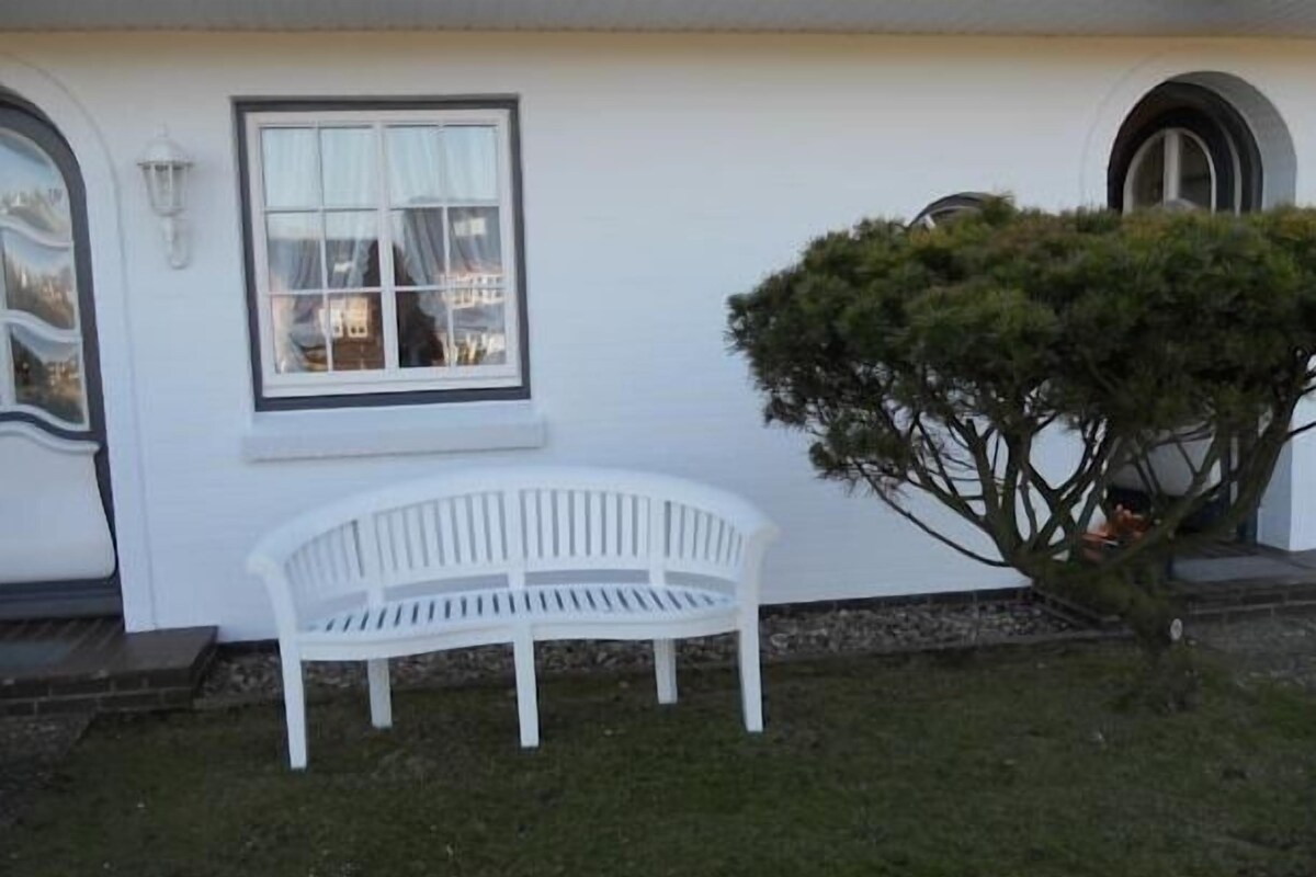 A white bench in front of a building with a window and a small tree.