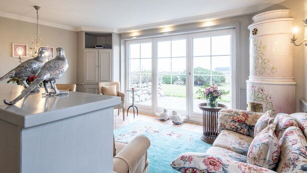 A spacious living room with a large window, a decorative metal sculpture, a floral sofa, and a white cabinet. - Ferienwohnung Sönshörn 8 Rechts (Sylt)