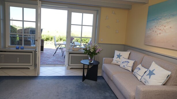 Ein Wohnzimmer mit einem beigefarbenen Sofa, einem kleinen schwarzen Beistelltisch mit einem Blumenstrauß und einem großen Fenster mit Blick ins Freie.