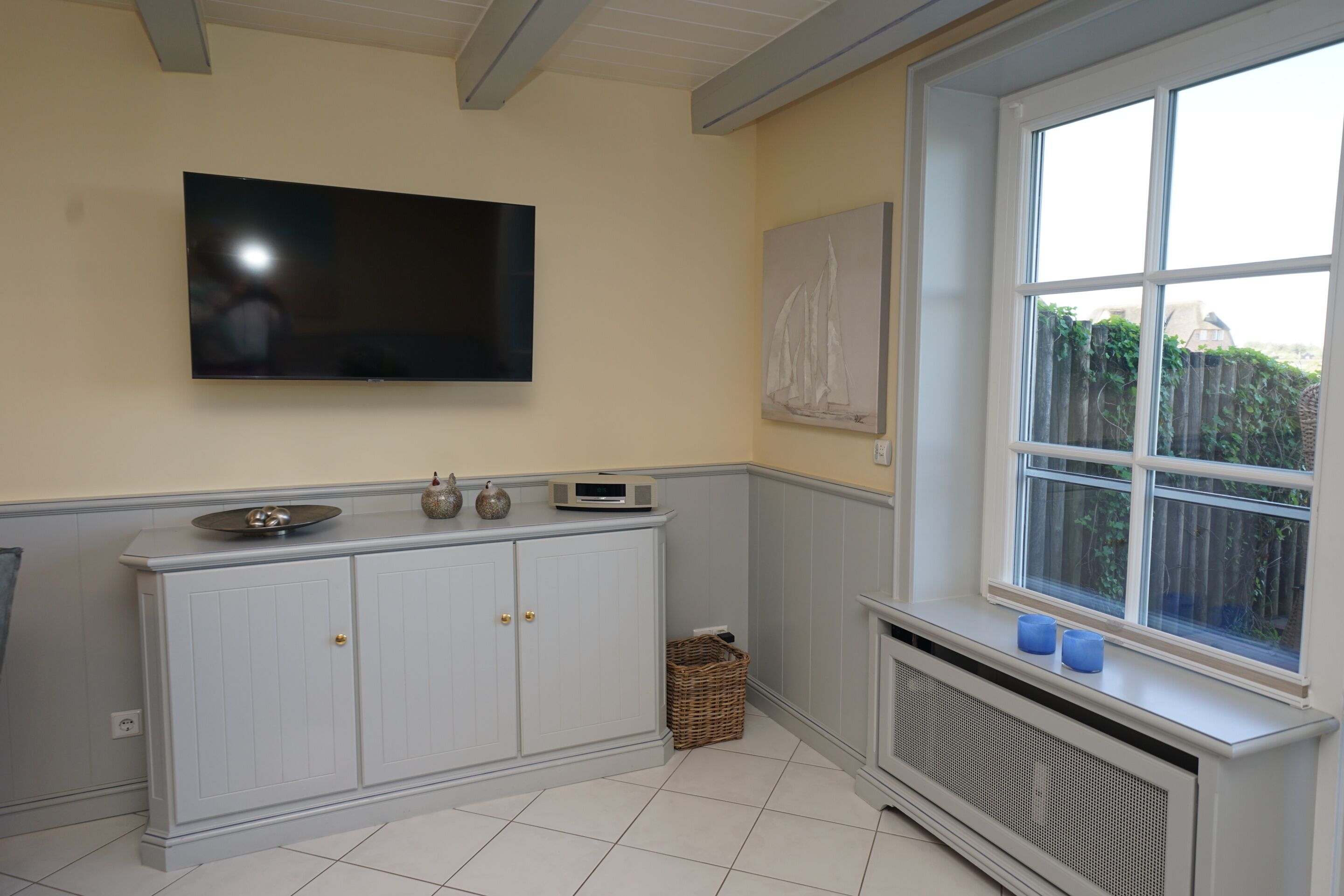 A kitchen with a flat-screen TV mounted on the wall, a white cabinet with two doors, a wicker basket, and a window with a view of greenery outside.
