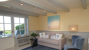 Ein Wohnzimmer mit einem beigen Sofa, einem blauen Sessel, einem kleinen Tisch und einem Fenster mit weißen Vorhängen.