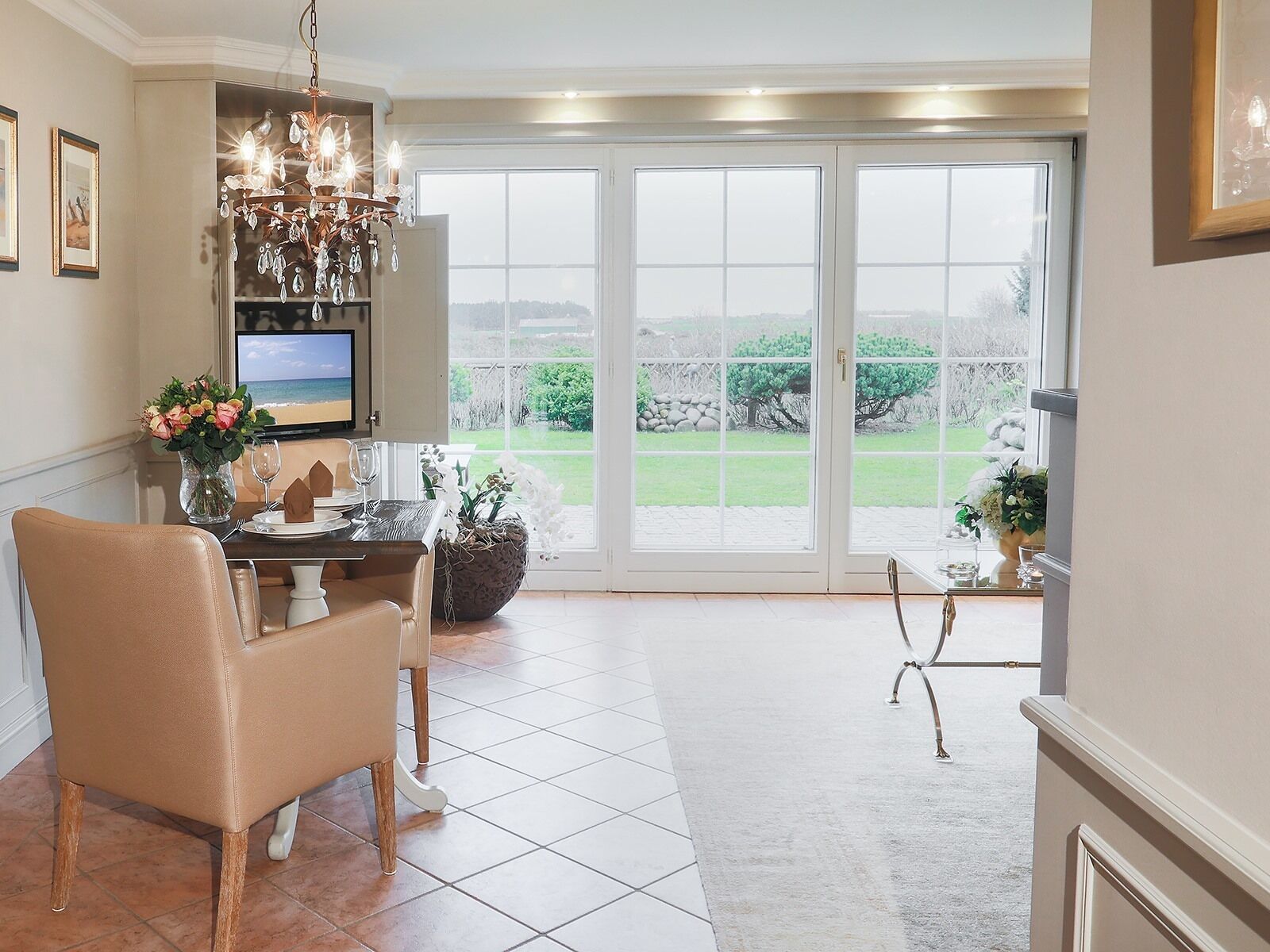 A dining area with a table set for two, a chandelier, and a view of the outdoors through large windows.