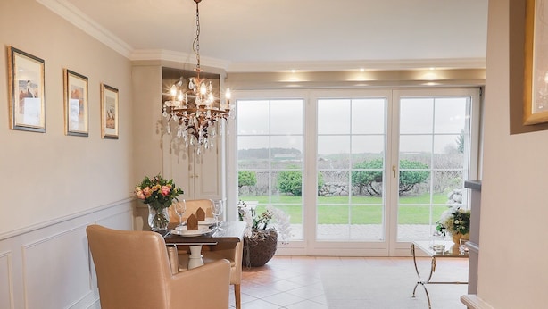 A dining area with a table set for four, a chandelier, and a view of a garden through large windows. - Ferienwohnung Sönshörn 8 Mitte (Sylt)