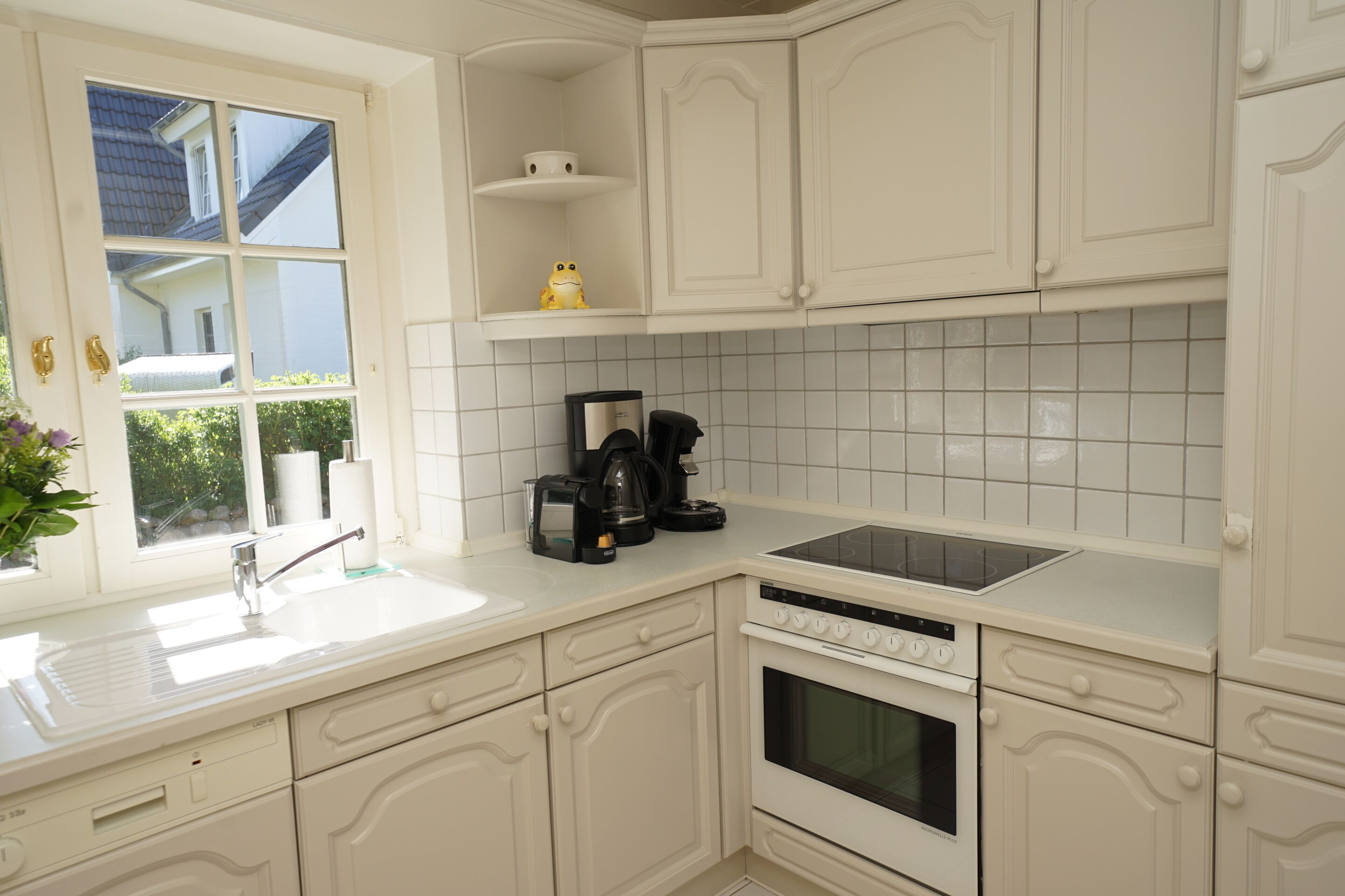 A kitchen with white cabinets, a built-in oven, a dishwasher, a coffee maker, and a window with a view of a house and greenery.