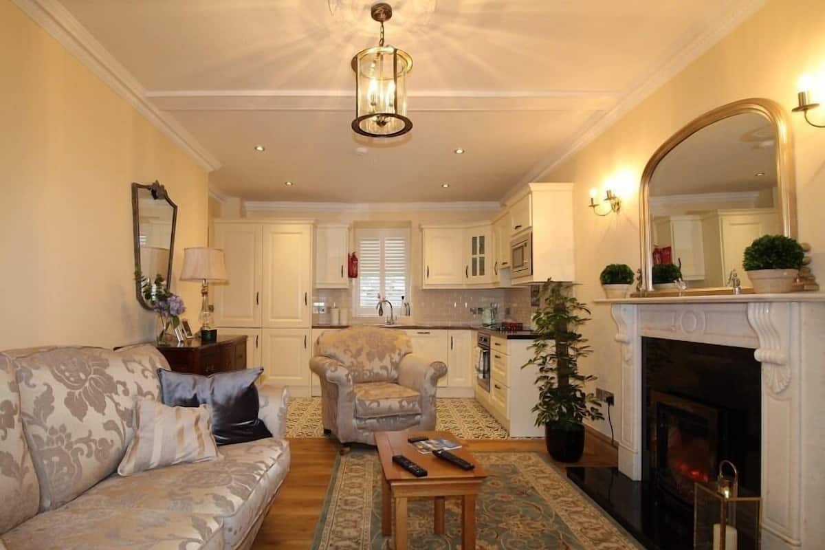 Een woonkamer met een bank, fauteuil en een open haard. Achtergrond toont een keuken met kasten en een raam.