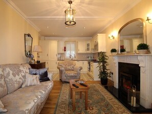 A living room with a sofa, armchair, and a fireplace. There is a kitchen in the background with cabinets and a window. - The Post House (Kinsale)