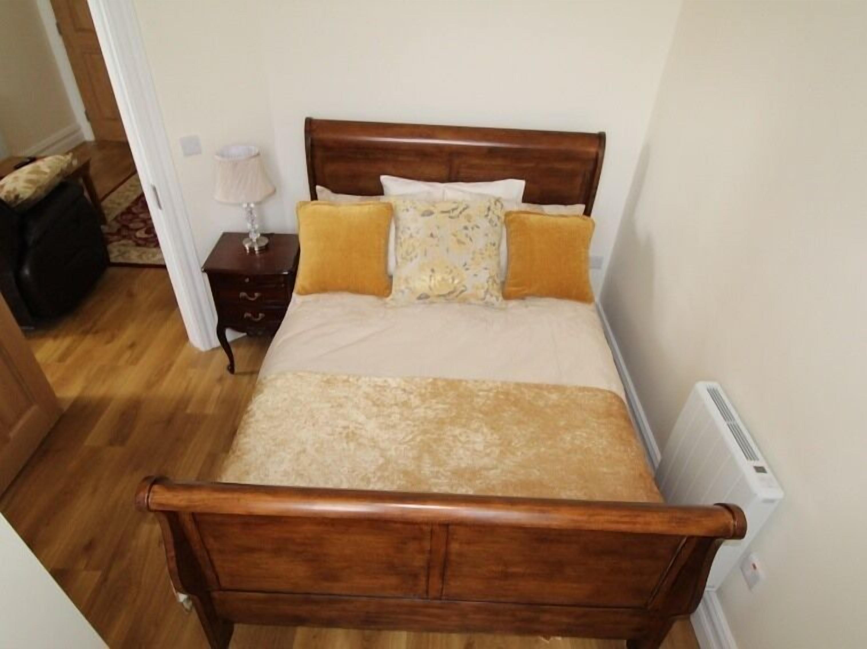 Una cama de madera con una colcha color beige y amarillo, una mesita de noche con una lámpara, y un radiador en la pared.