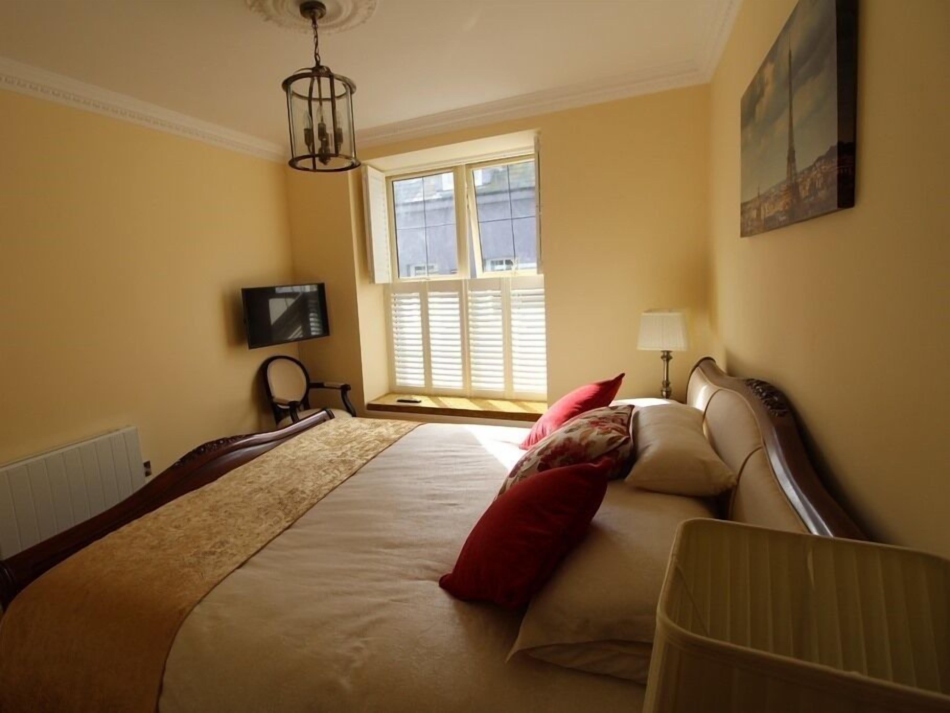 A hotel room with a large bed, a television on the wall, a window with blinds, and a painting above the bed.