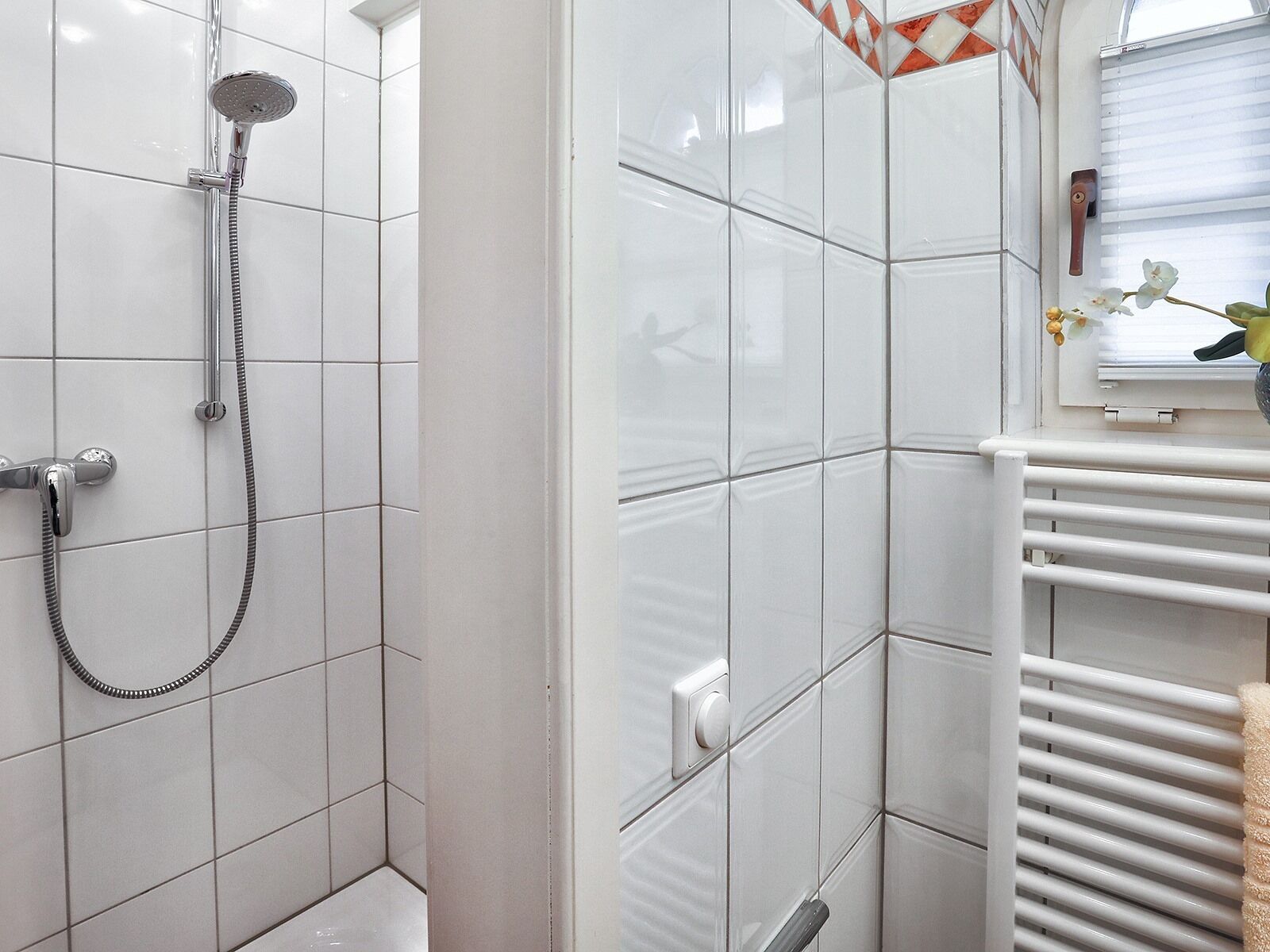 Ein Badezimmer mit weißen Fliesen, einer Dusche inklusive Duschbrause, einem Handtuchhalter und einem Fenster mit Blick nach draußen.