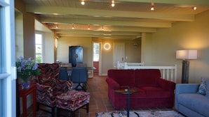 Un vaste espace de vie comprenant un canapé rouge, un fauteuil bleu et un fauteuil à motifs. Une petite table ronde avec une lampe se trouve au centre de la pièce. Le sol est carrelé et le plafond est orné de poutres apparentes et de luminaires encastrés.