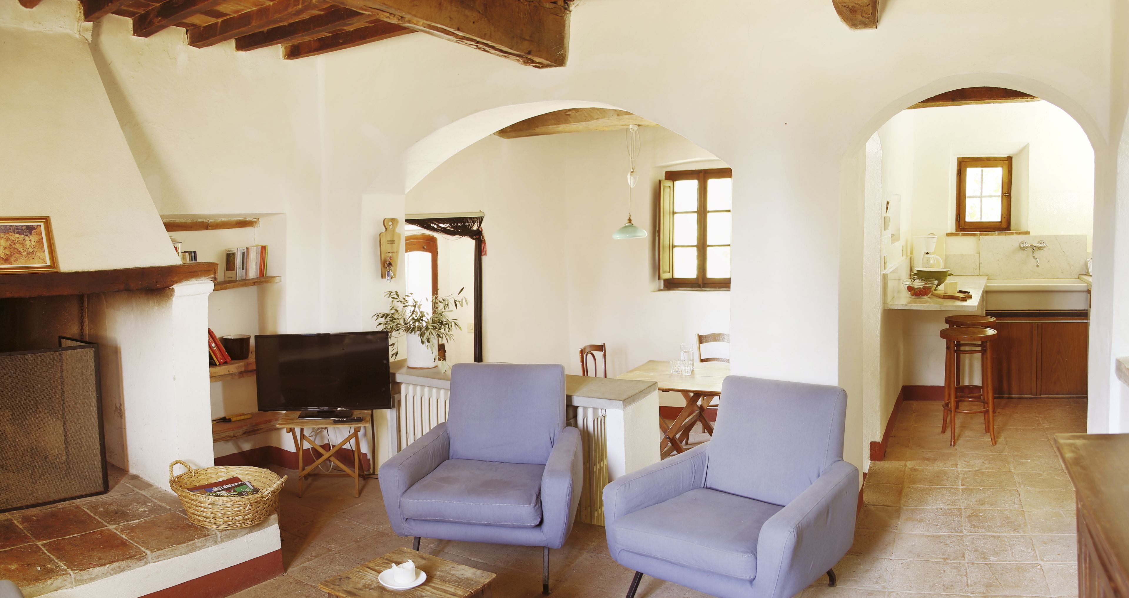 A living room with two blue armchairs, a television, and a fireplace. There is a kitchen visible through an archway.