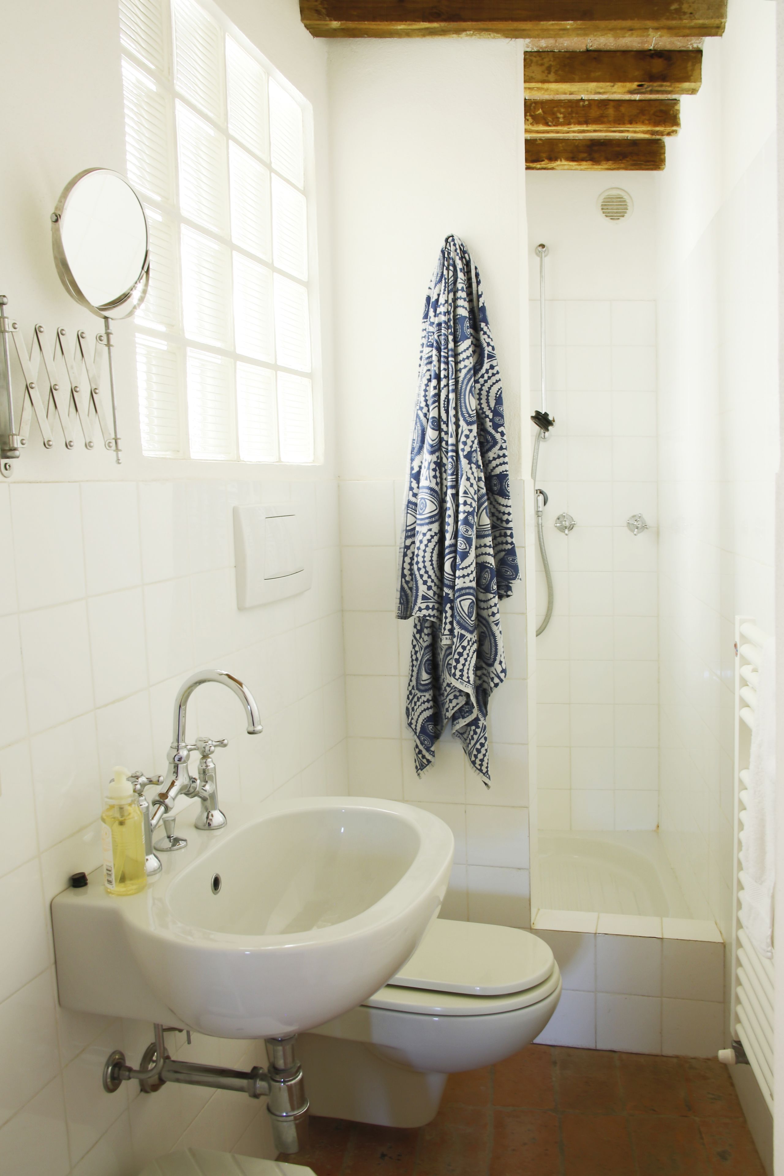 Baño con lavamanos, inodoro y ducha blancos. Hay una toalla colgada en la pared y una ventana con persianas.