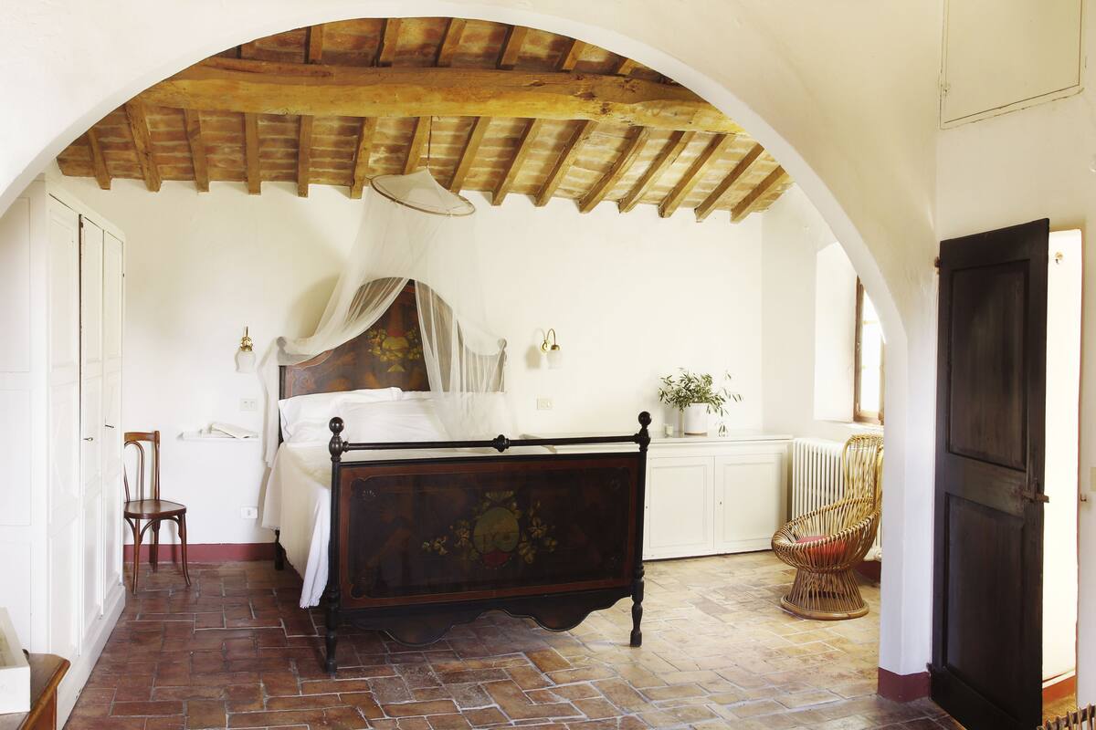 A bedroom with a four-poster bed, a wooden ceiling, a white wall, a window, and a wicker chair.