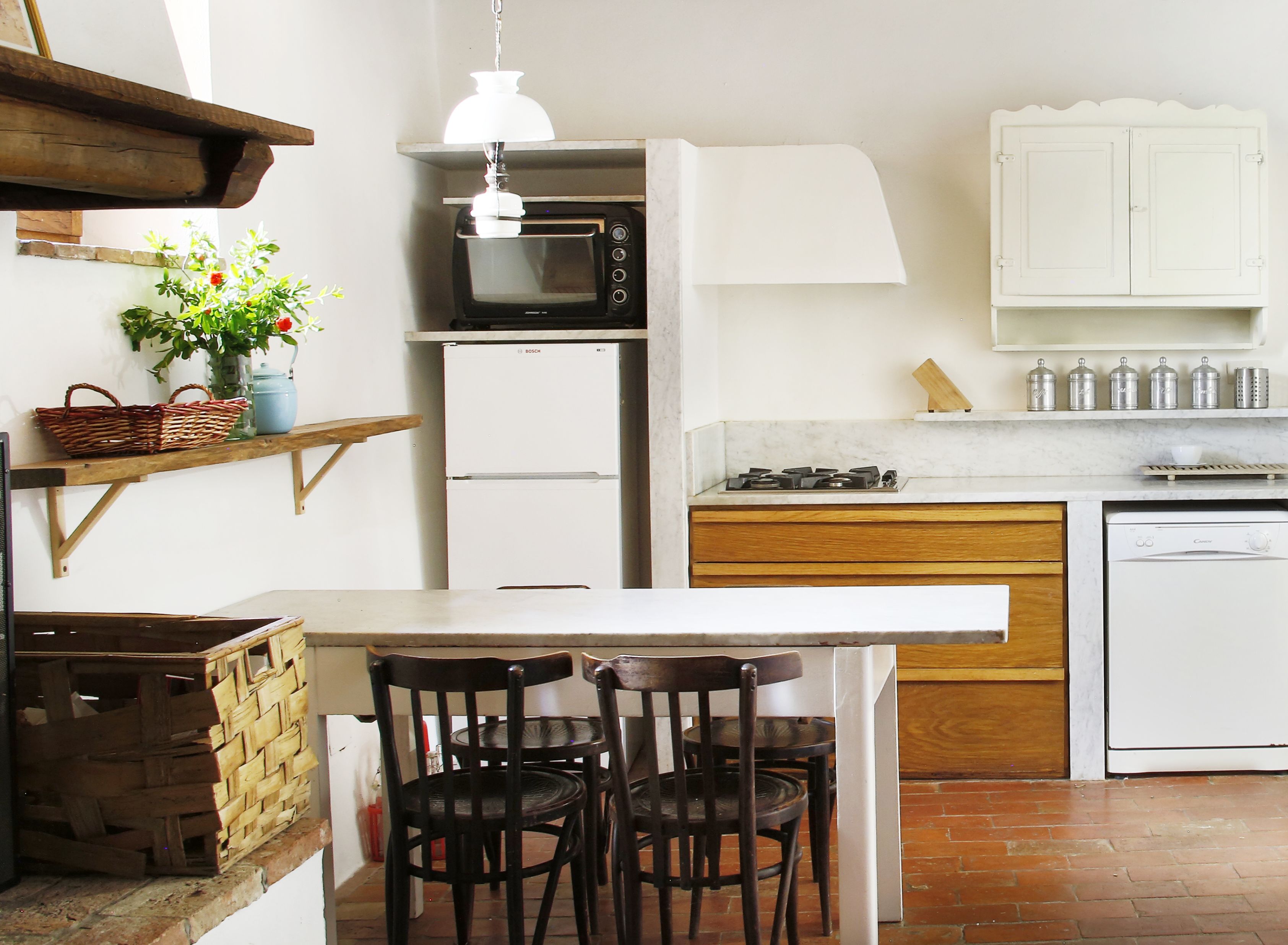 Una cocina compacta equipada con microondas, refrigerador y lavavajillas. Hay una mesa de madera con sillas, una lámpara colgante y un estante con una cesta y flores.