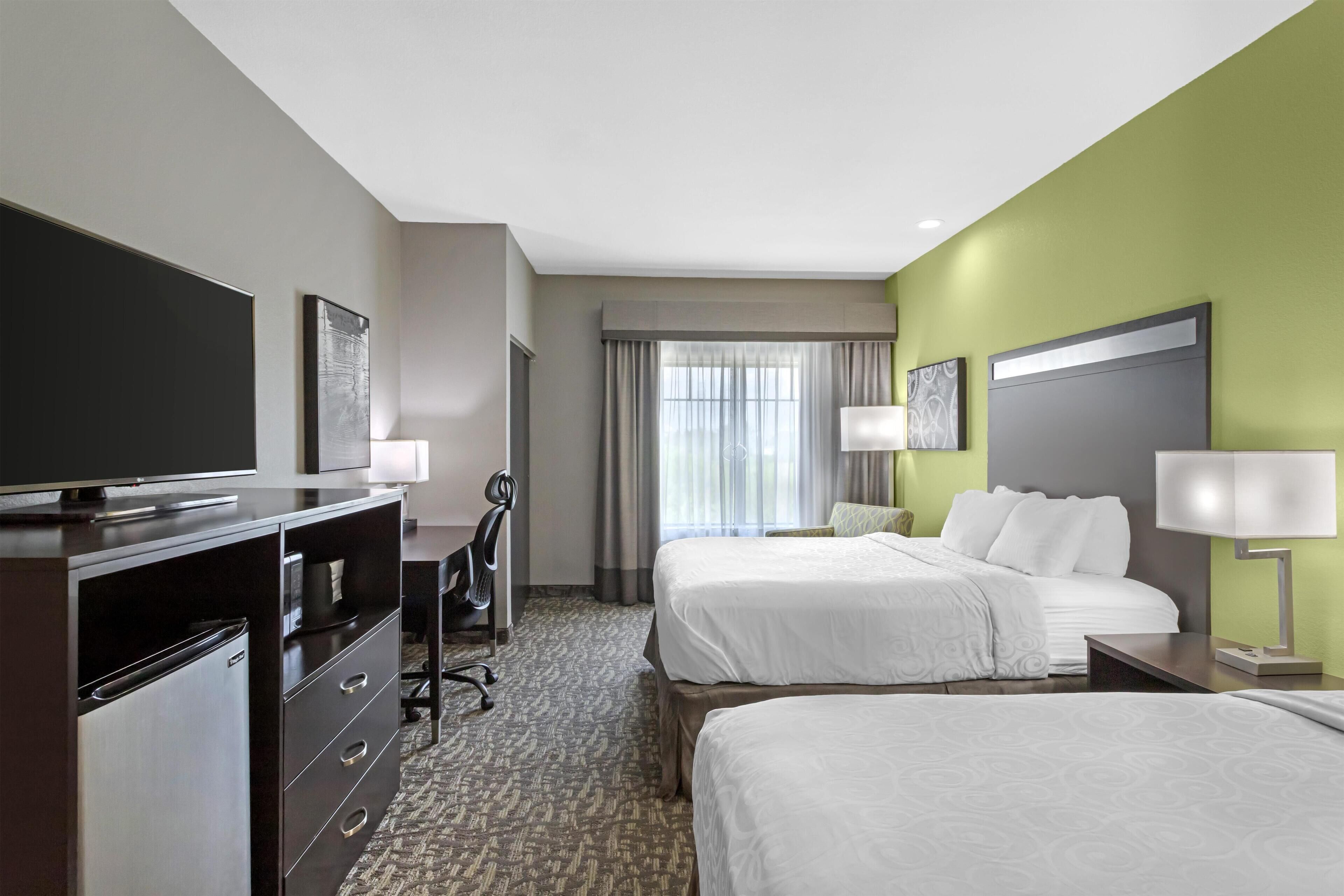 A hotel room with two beds, a desk, a television, and a large window with curtains.