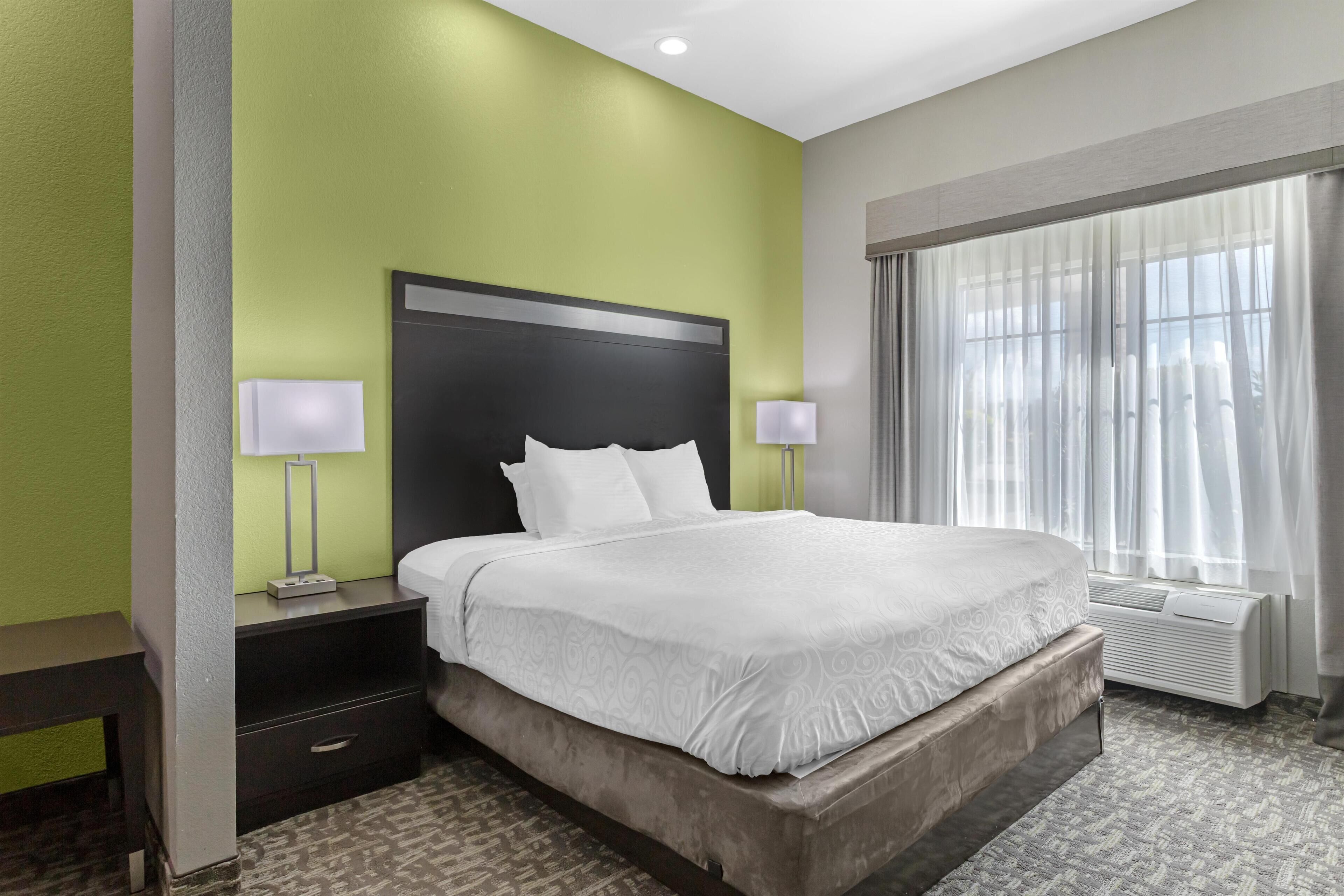 Una habitación de hotel con una cama grande, una mesita de noche con una lámpara, una ventana con cortinas translúcidas y un calefactor en la pared.