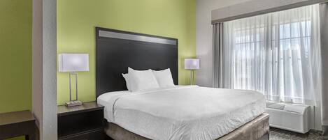 Una habitación de hotel con una cama grande, una mesita de noche con una lámpara, una ventana con cortinas translúcidas y un calefactor en la pared.