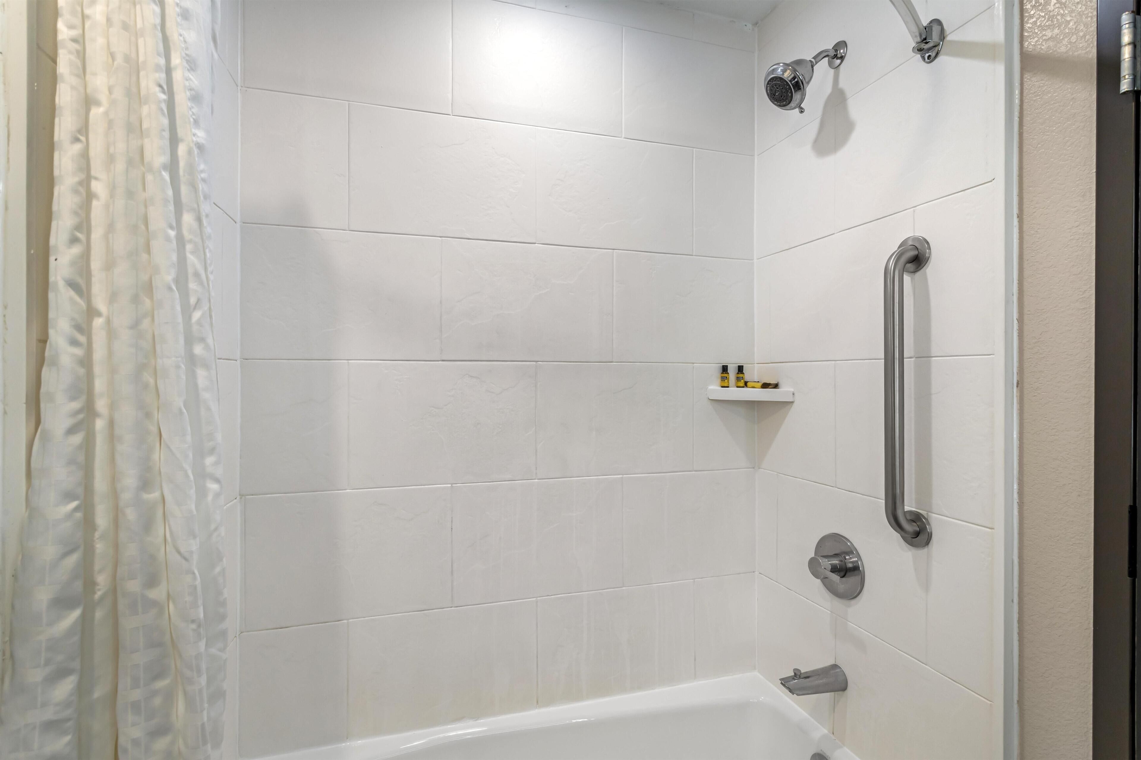 Un baño moderno con azulejos blancos, una ducha con cabezal de ducha manual, una barra de apoyo y un estante con artículos de aseo.