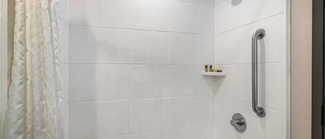 Un baño moderno con azulejos blancos, una ducha con cabezal de ducha manual, una barra de apoyo y un estante con artículos de aseo.