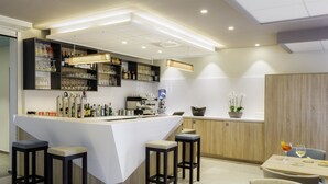 Zona de bar moderna amb un taulell blanc, tamborets, prestatges amb copes i ampolles, una màquina de cafè i una taula amb una beguda i un plat.