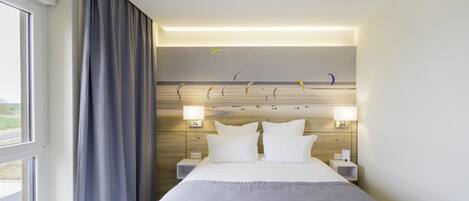 Habitación de hotel con una cama grande, dos mesitas de noche, cabecera con luces empotradas y una ventana con cortinas.